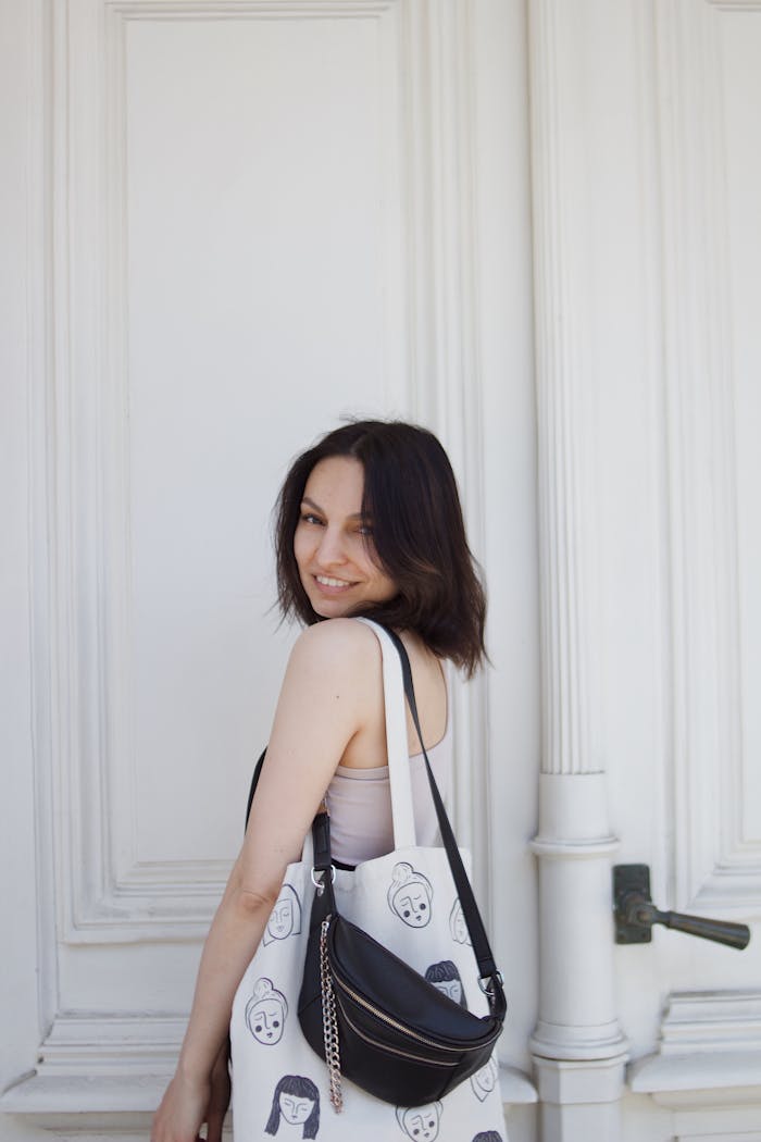 Smiling woman posing with a fashionable tote and sling bag against a white wall.
