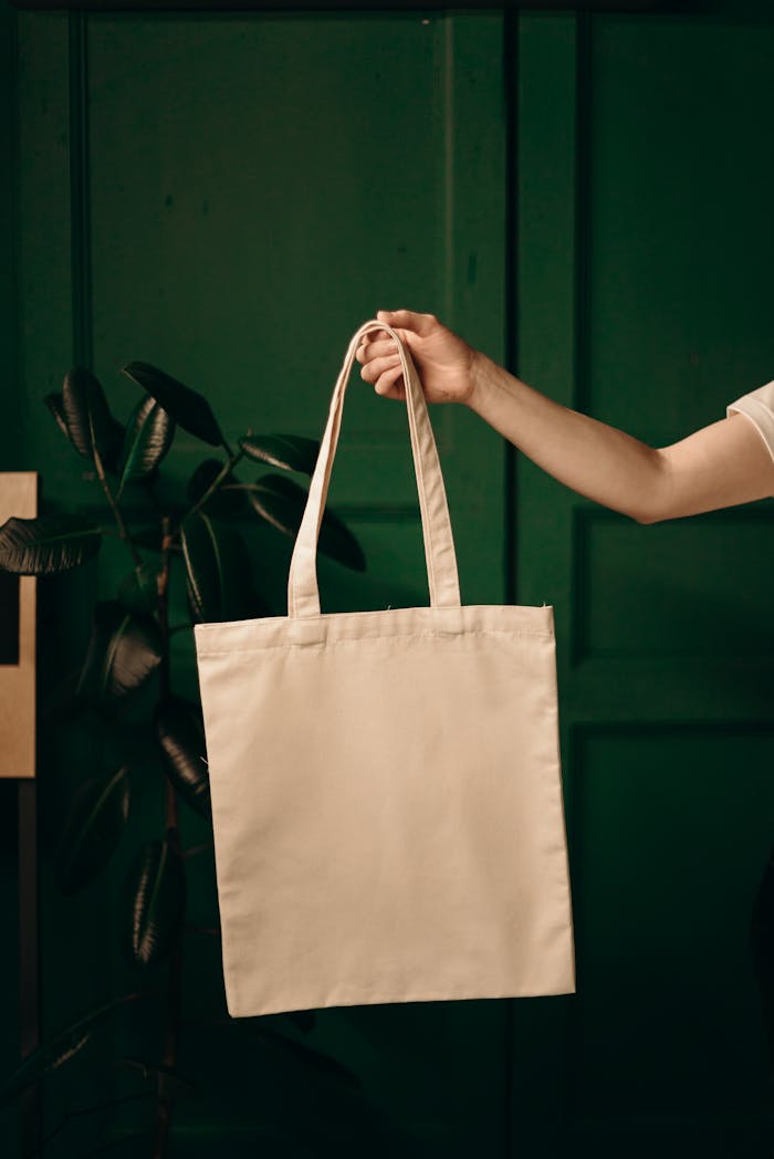 Simple cotton tote bag held by a hand against a green background, ideal for eco themes.