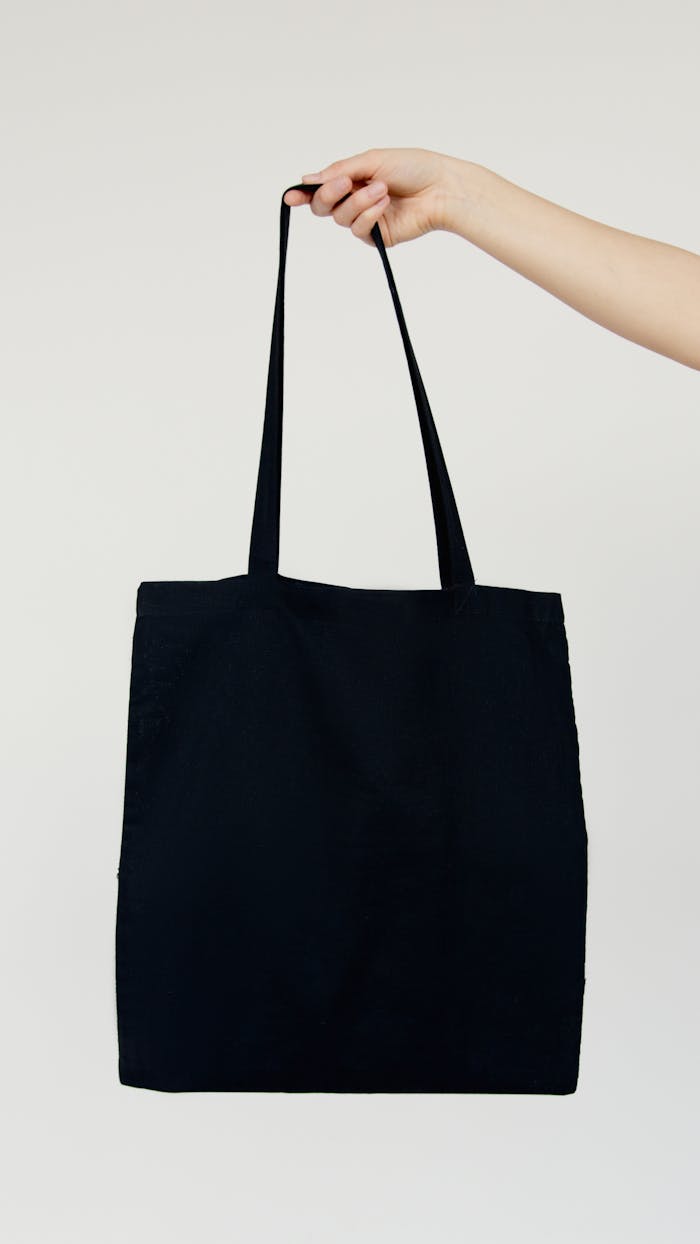 Minimalist image of a hand holding a blank black tote bag against a neutral background.